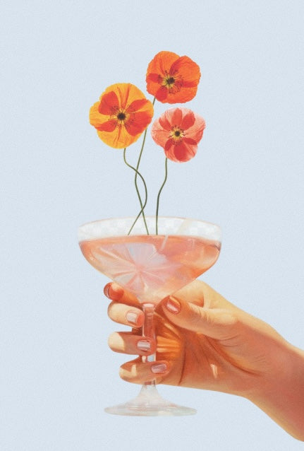 Hand holding a coupe glass filled with a light pink drink, garnished with three vibrant poppy flowers in shades of orange, yellow, and pink, against a pale blue background.