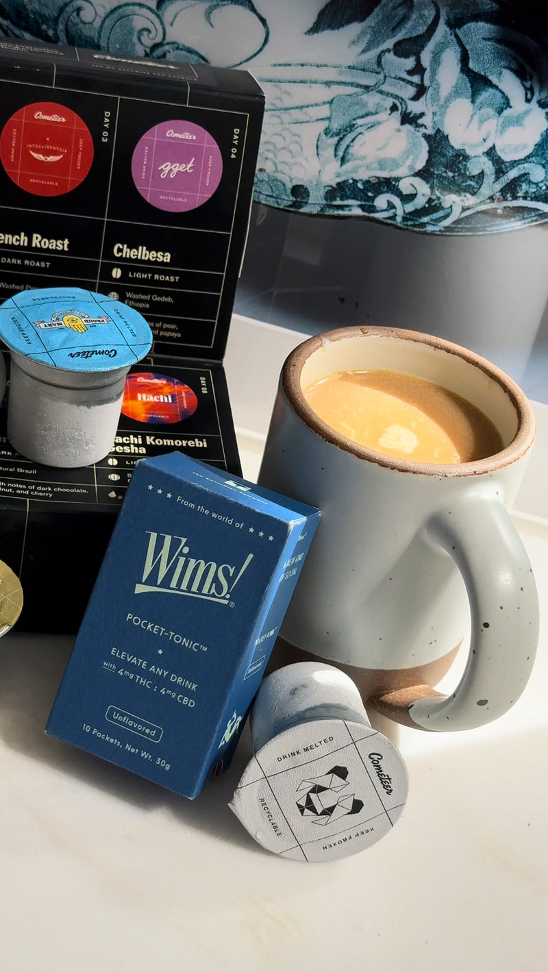 A cozy setup featuring a steaming mug of creamy hot latte alongside a blue Wims Unflavored Pocket-Tonic™ box, labeled “Elevate Any Drink” with 4mg THC and 4mg CBD. Next to the box is a Cometeer coffee capsule, with additional capsules and a black Cometeer coffee box in the background. The scene evokes warmth and comfort, highlighting the perfect ingredients for a soothing THC/CBD coffee drink.