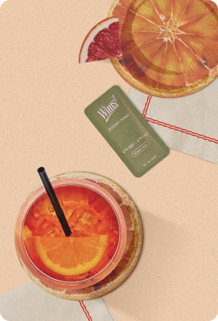 A top-down view of a vibrant orange mocktail in a clear glass filled with ice and garnished with a fresh orange slice. The drink sits on a round coaster beside a Wims Pocket-Tonic packet, with sliced citrus and a folded napkin on a warm-toned surface.