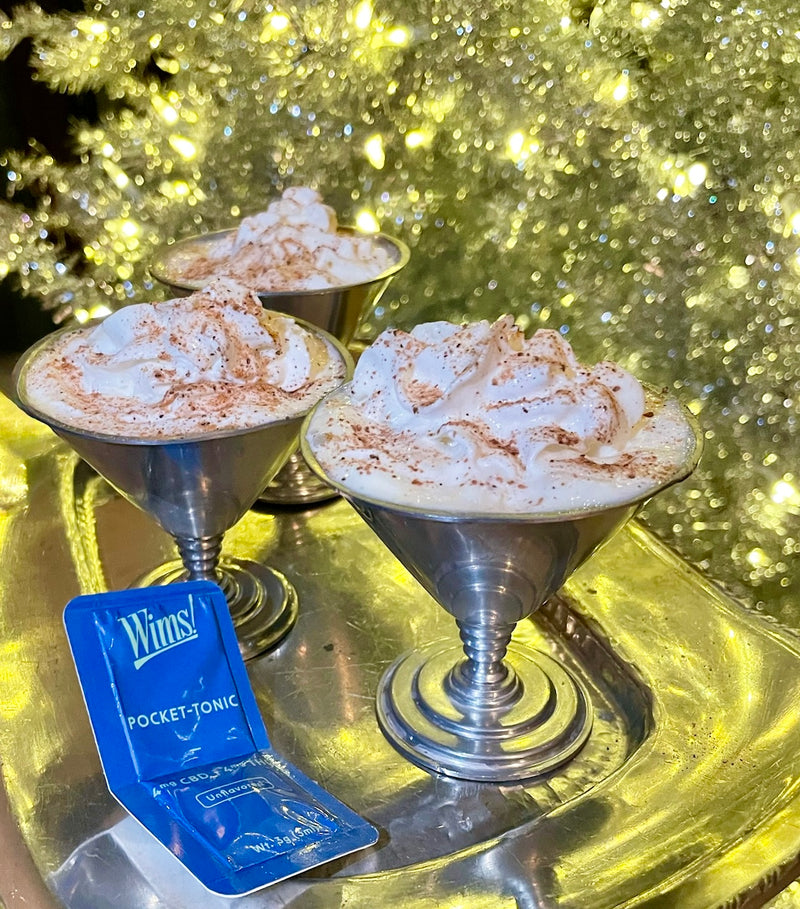 Three Egg Nog Shooters in silver glasses, topped with whipped cream and a sprinkle of nutmeg, served on a shiny tray with a Wims Pocket-Tonic™ (4mg THC, 4mg CBD) sachet, glowing Christmas tree lights in the background.