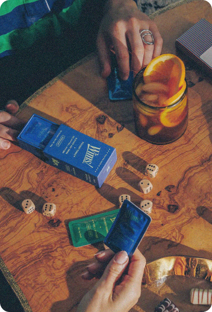 Close-up of two people playing a dice game at a wooden table with a glass of iced citrus drink and various game pieces. One person opens a blue Wims Pocket-Tonic box while another holds a packet. Decorative items and small treats sit nearby, creating a cozy, playful atmosphere.