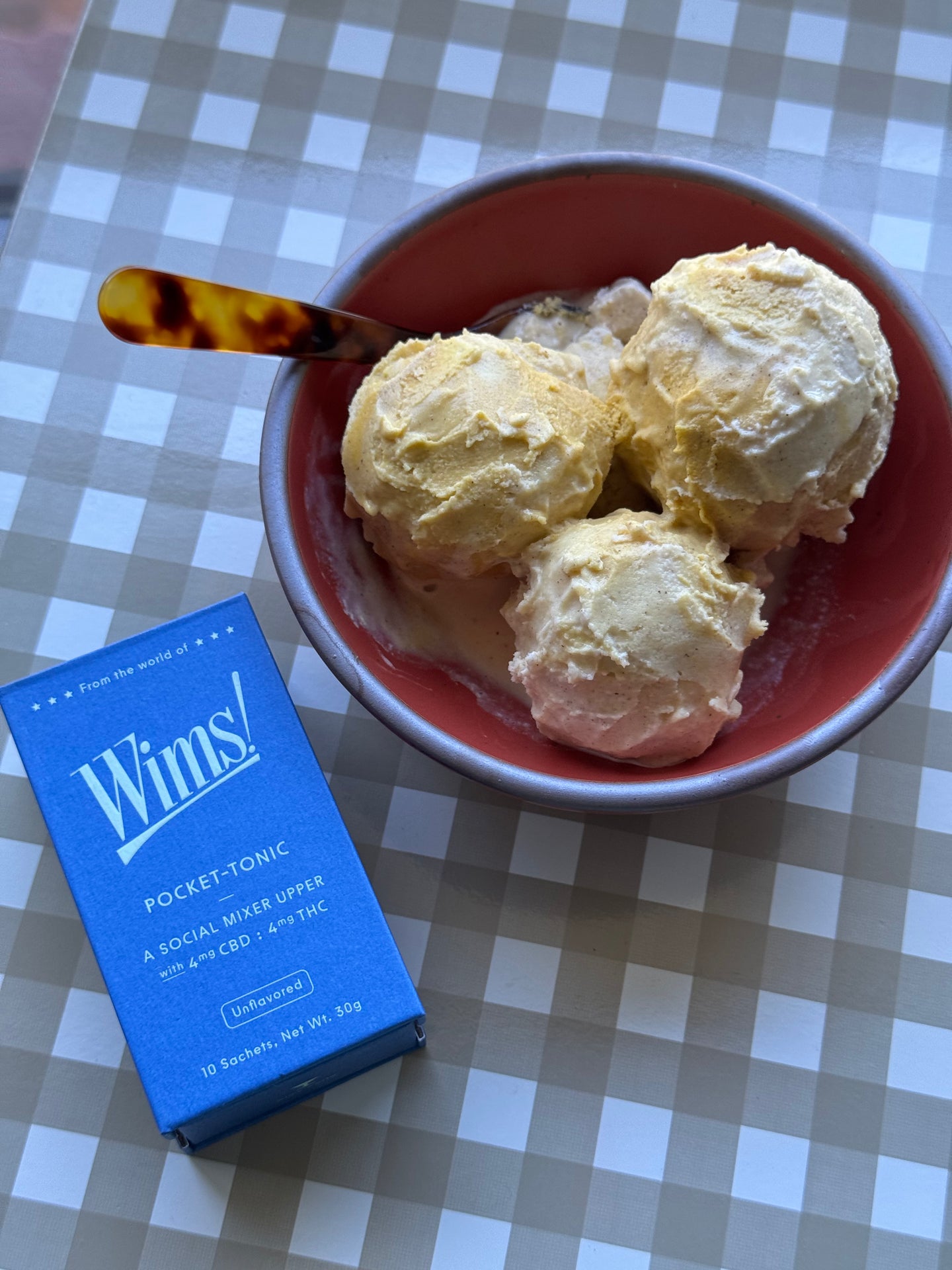Bowl of homemade pumpkin pie ice cream with three scoops served in a coral dish, next to a Wims Unflavored Pocket-Tonic™ (4mg THC, 4mg CBD) box on a checkered table surface.