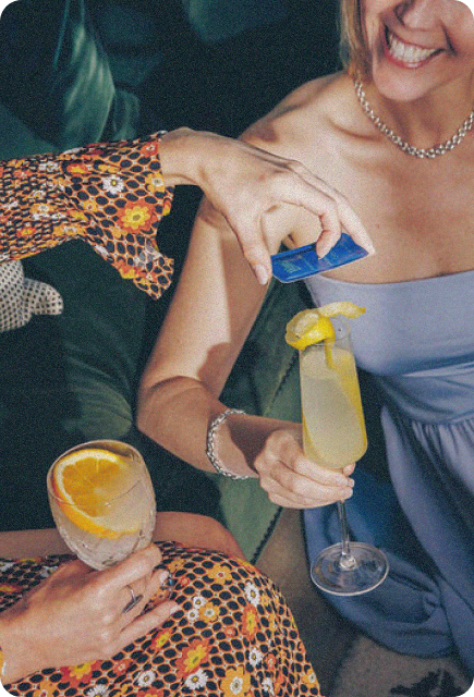 Two women sit together on a couch, smiling and holding cocktails garnished with citrus slices. One woman pours a blue Wims Pocket-Tonic into the other’s drink, creating a cheerful, social moment in a relaxed setting.
