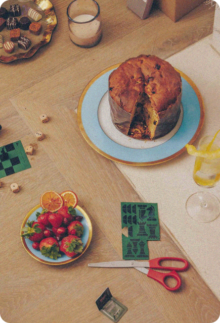 A tabletop scene with holiday treats and crafting materials: a sliced panettone sits on a blue and gold plate, a small dish of strawberries and citrus slices is nearby, and red-handled scissors lie next to green cut-out game pieces. Dice, chocolates, and a glass with a lemon twist add to the cozy, festive setting.