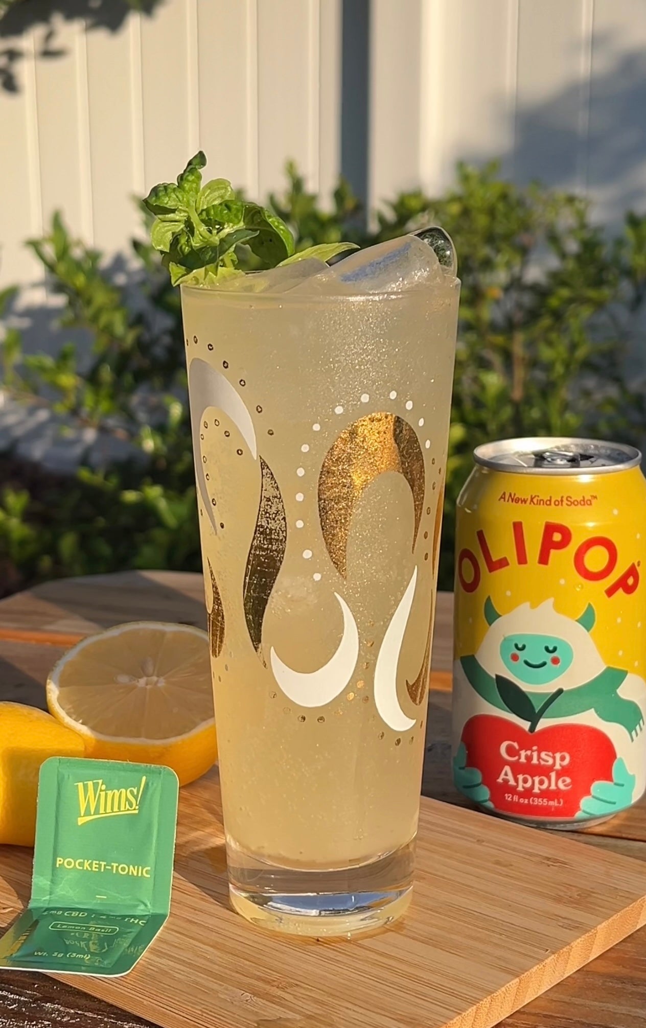 A tall glass of Sunny Cider Spritz mocktail filled with ice and garnished with a fresh basil sprig. The drink has a light golden hue, with visible bubbles, sitting on a wooden board. Surrounding it are a halved lemon, a green Wims Lemon Basil Pocket-Tonic™ sachet, and a can of Olipop Crisp Apple, creating a cozy, autumn-inspired setting for this THC/CBD fall drink.