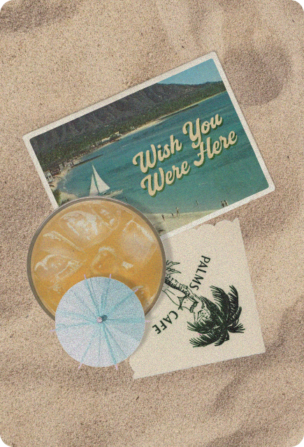 A vintage beach postcard that reads “Wish You Were Here” lies on sand next to a cocktail glass with ice and a small paper umbrella, along with another postcard featuring a palm tree illustration.