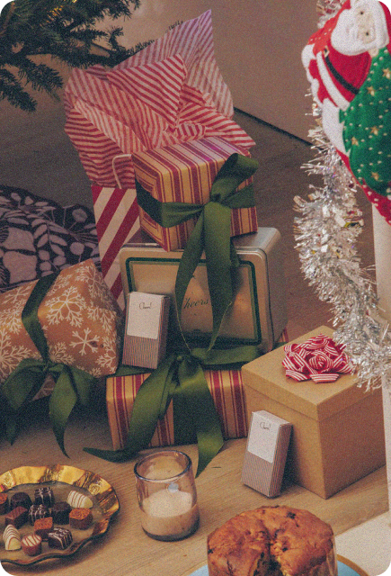 Wrapped holiday gifts with green ribbons and candy-stripe wrapping sit under a Christmas tree, with small Wims Pocket-Tonic™ packets placed among them. A tinsel garland, chocolates, a candle, and a loaf cake create a cozy, festive gifting scene.