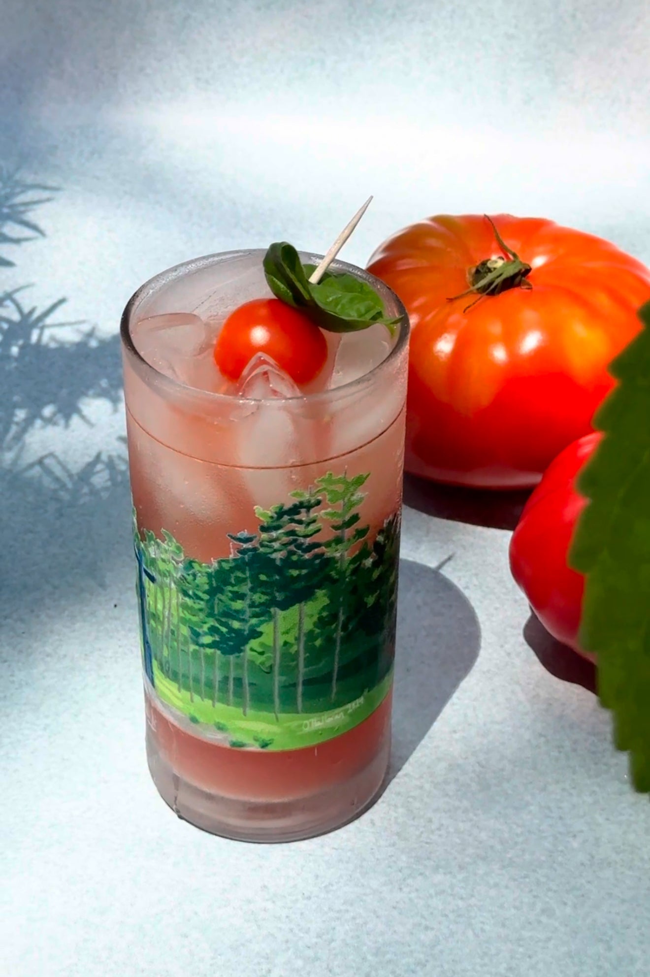 A Collins glass decorated with a green tree illustration, filled with a pinkish-red tomato spritz over ice. The drink is garnished with a cherry tomato and fresh basil leaf skewered on a toothpick. Two ripe tomatoes rest in the background on a light blue surface, with soft leaf shadows falling across the scene.