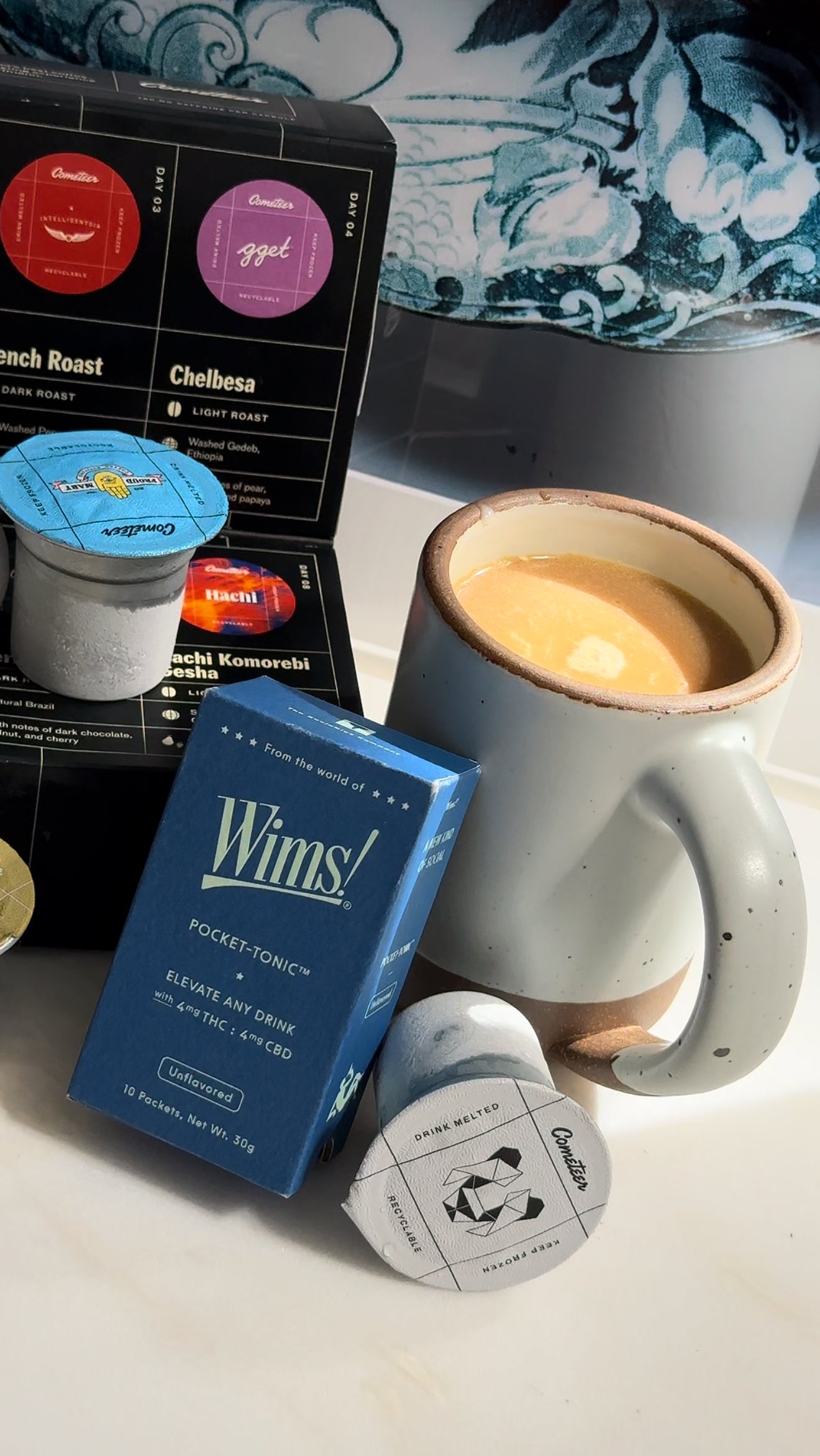A cozy setup featuring a steaming mug of creamy hot latte alongside a blue Wims Unflavored Pocket-Tonic™ box, labeled “Elevate Any Drink” with 4mg THC and 4mg CBD. Next to the box is a Cometeer coffee capsule, with additional capsules and a black Cometeer coffee box in the background. The scene evokes warmth and comfort, highlighting the perfect ingredients for a soothing THC/CBD coffee drink.