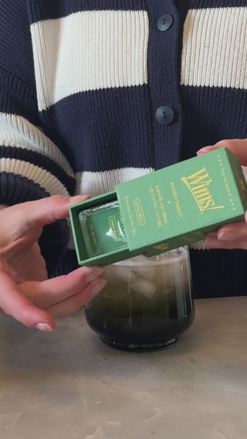 A rounded rocks glass with an iced tea and lemonade mixture is shown as a Lemon Basil Wims Pocket-Tonic™ is snapped open into the drink. A fresh lemon wedge is placed on the rim as the final garnish.