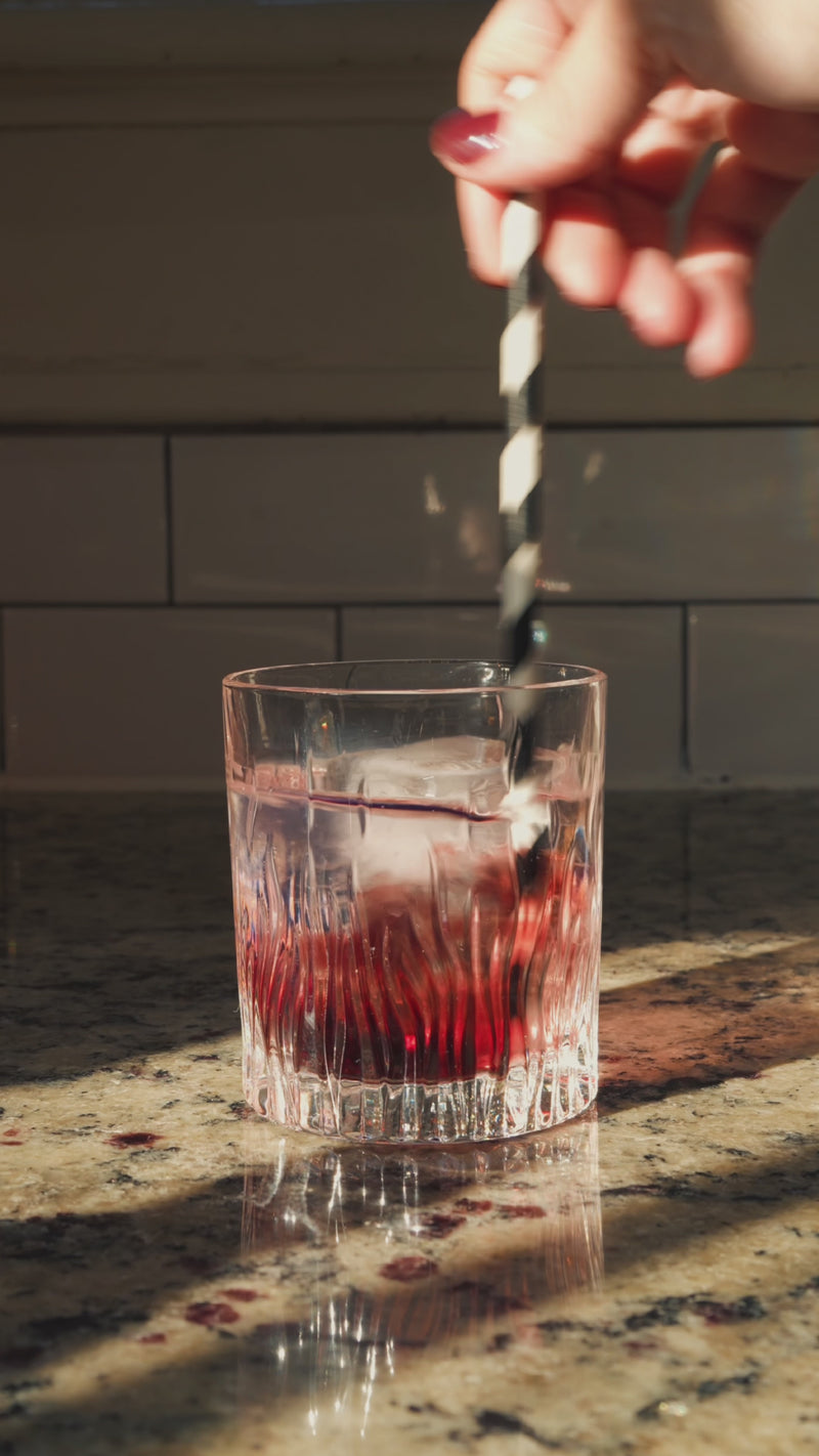 A short rocks glass is shown as a Lemon Basil Wims Pocket-Tonic™ is snapped open directly into the drink, then the mixture is gently stirred with a straw.
