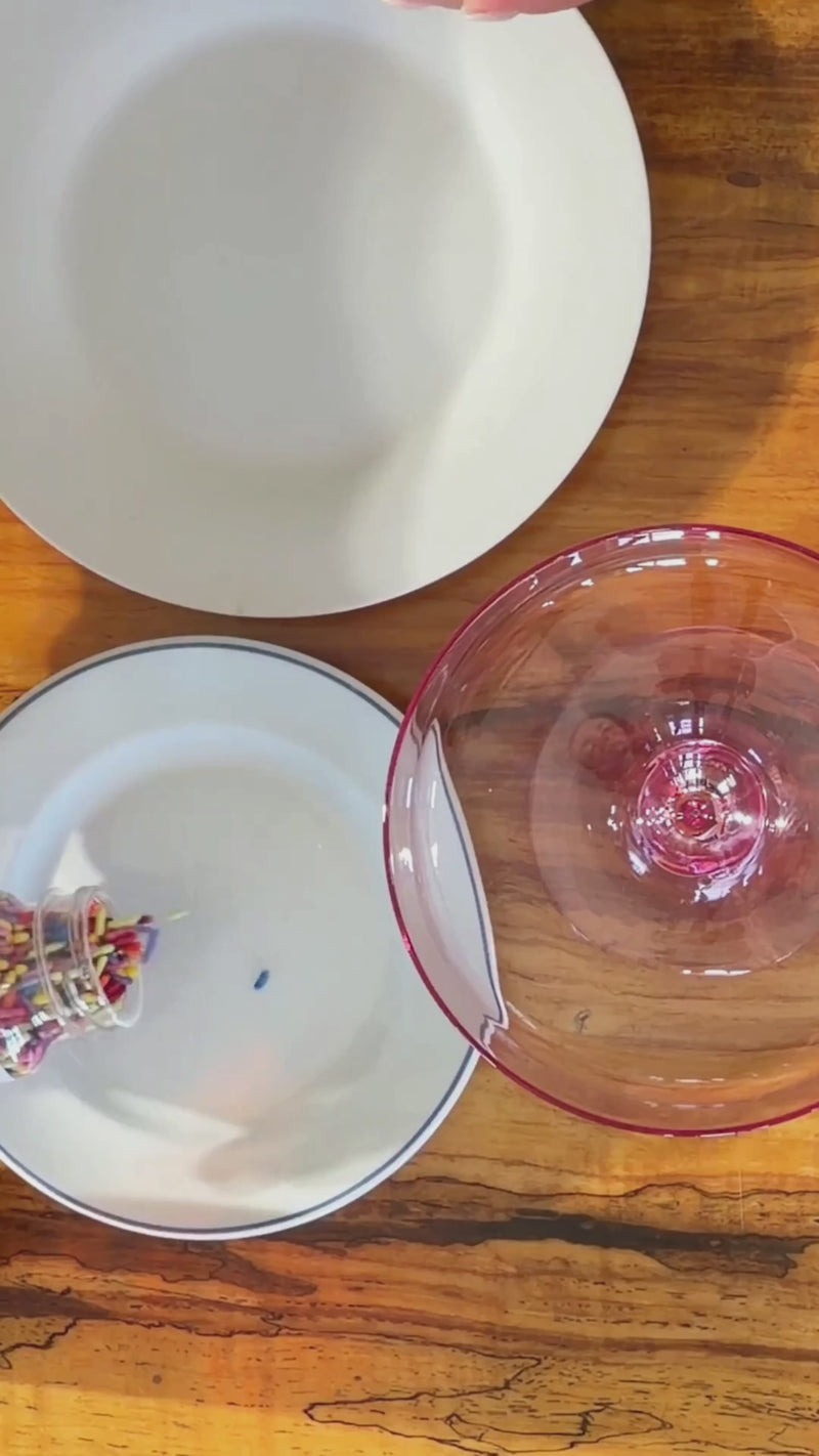 Agave nectar being poured onto a plate beside rainbow sprinkles as the rim of a cocktail glass is coated and dipped to create a colorful rainbow garnish.