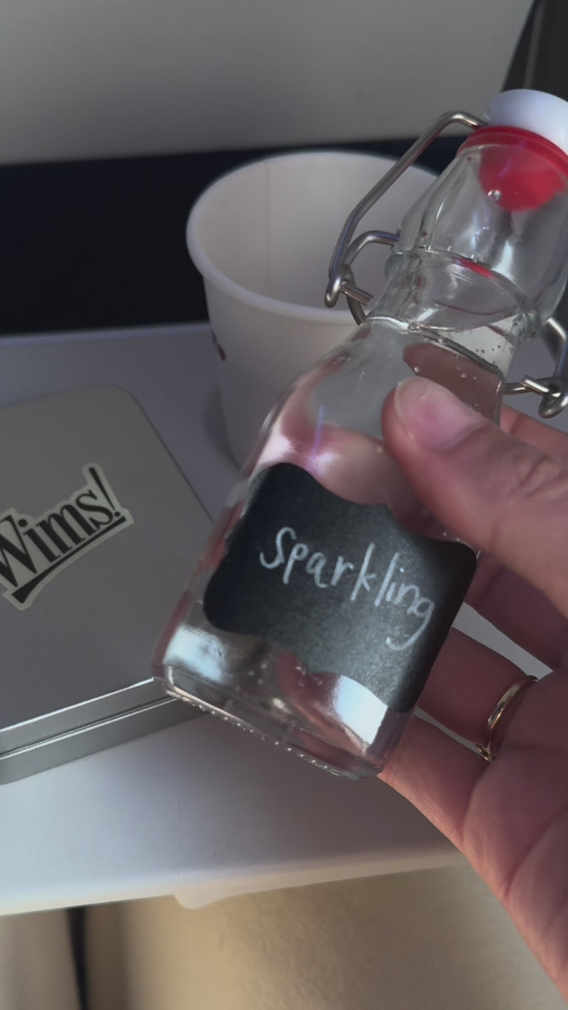 A clear jar labeled “Sparkling” pours pineapple juice into a white cup on an airplane tray table.
