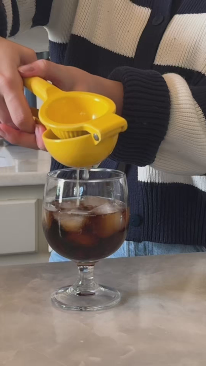 A short stemmed glass filled with cola and ice is shown as a fresh lime wedge is squeezed over the top, with citrus juice dripping into the drink to brighten the flavor.
