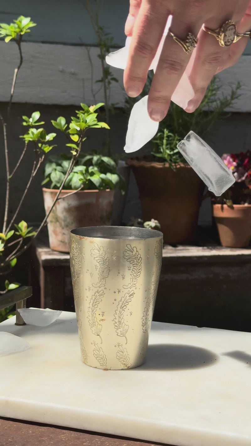 Close-up of a silver mixing cup with leaf details, filled with ice, as tequila and rosemary thyme syrup are poured in. Green plants fill the background, and only a hand is visible.