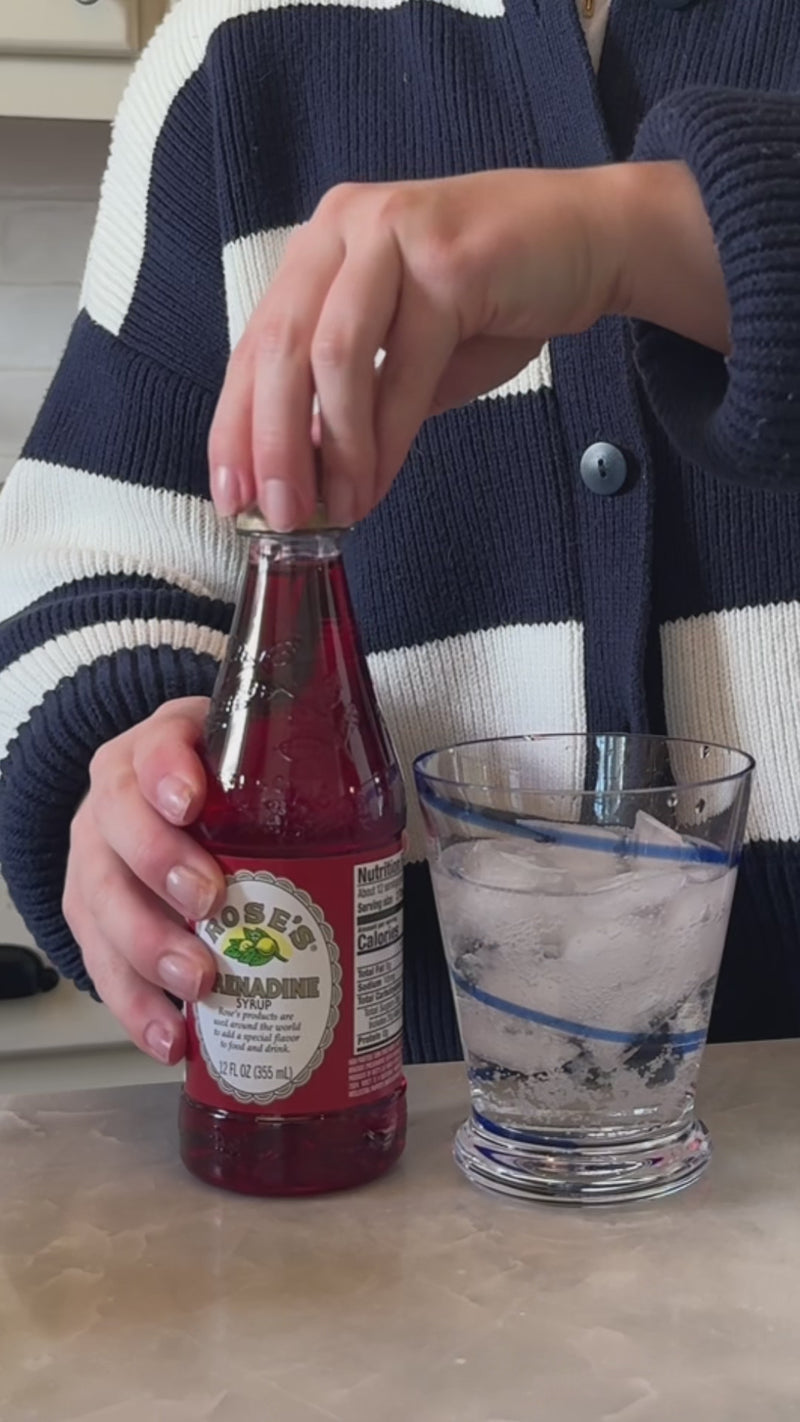 A rocks glass filled with bubbly citrus soda is shown as bright red grenadine is poured in, slowly settling toward the bottom and creating a layered cherry glow through the ice.