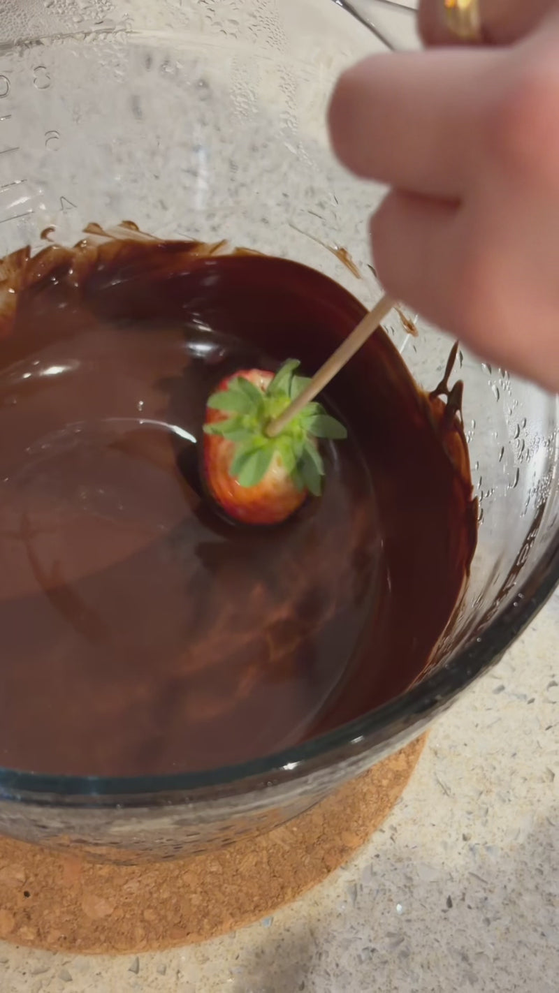 Fresh strawberry being dipped into melted dark chocolate in a glass bowl on a kitchen countertop for even coating.