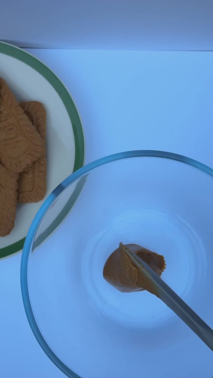 A clear glass mixing bowl on a white surface while a mixer blends sweetened condensed milk, whipping cream, cookie butter, and cinnamon until smooth.