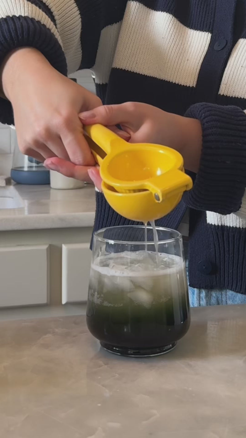 A rounded rocks glass filled with iced tea, lemonade, and ice is shown as a fresh lemon wedge is squeezed over the top, with citrus juice dripping into the drink.