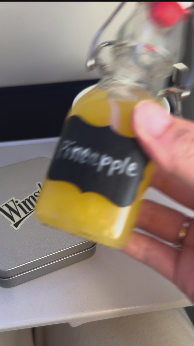 A clear jar labeled “Pineapple” pours pineapple juice into the same cup. A packet of sweetener is opened and poured into the drink.