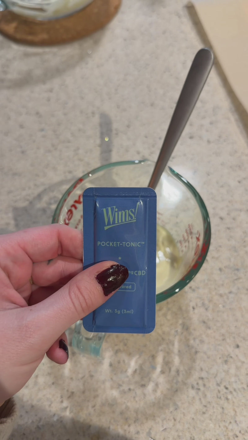 Unflavored Wims Pocket-Tonic™ being added to melted white chocolate in a glass measuring cup on a kitchen countertop before mixing.