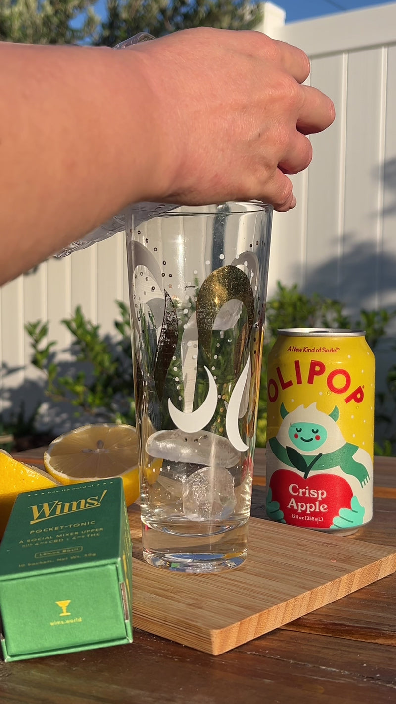 Filling a tall glass with ice, then pouring Olipop Crisp Apple over it to create a crisp and fruity base. Fresh lemon slices, a Wims Lemon Basil Pocket-Tonic™ box, and an Olipop Crisp Apple can sit nearby on a wooden board, evoking a cozy fall vibe.