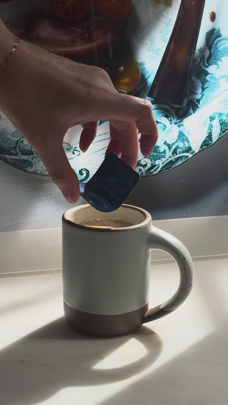 A hand snaps open a Wims Unflavored Pocket-Tonic™ sachet and pours it into the hot latte, then stirs gently. This step adds a subtle herbal twist that balances and enhances the rich, bold coffee flavor.