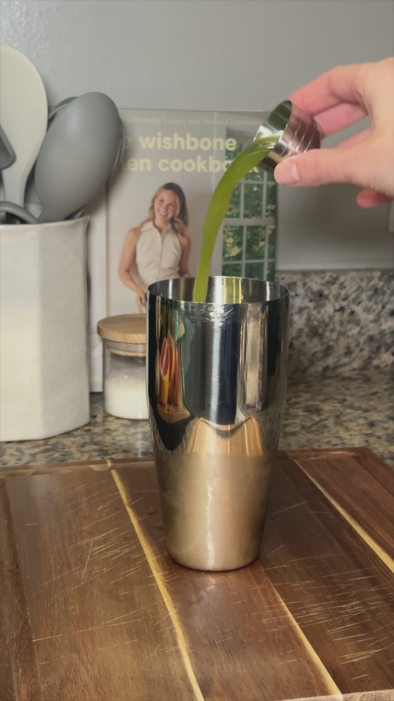 Pouring fresh green cucumber juice into a stainless steel cocktail shaker with simple syrup or agave, ice, and Wims Unflavored Pocket-Tonic™, set on a wooden cutting board with a kitchen counter, utensil holder, candle, and cookbook in the background.