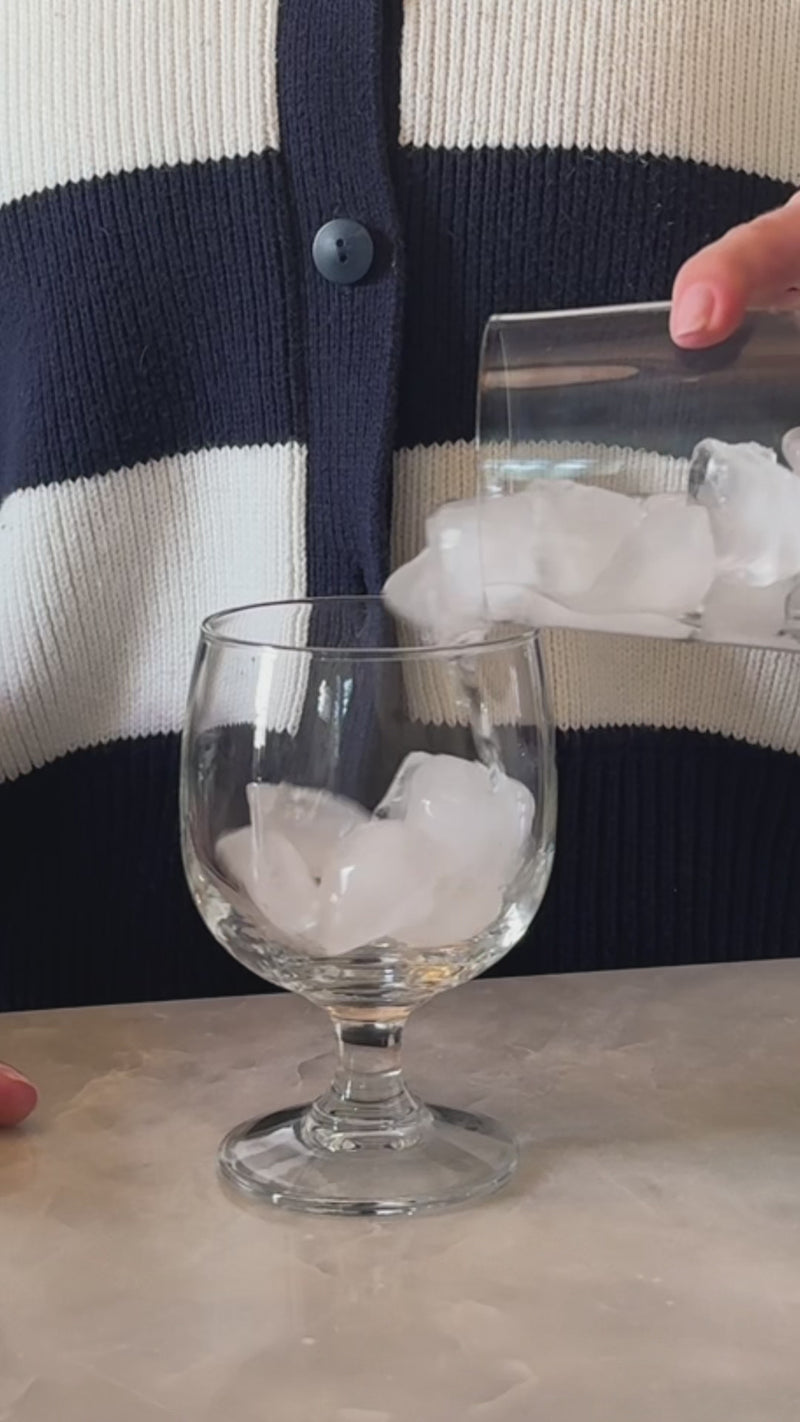 A clear, short stemmed glass with a rounded bowl sits on a neutral stone surface as ice fills the glass. Coca-Cola is poured over the ice, creating visible bubbles and a deep, fizzy cola base.