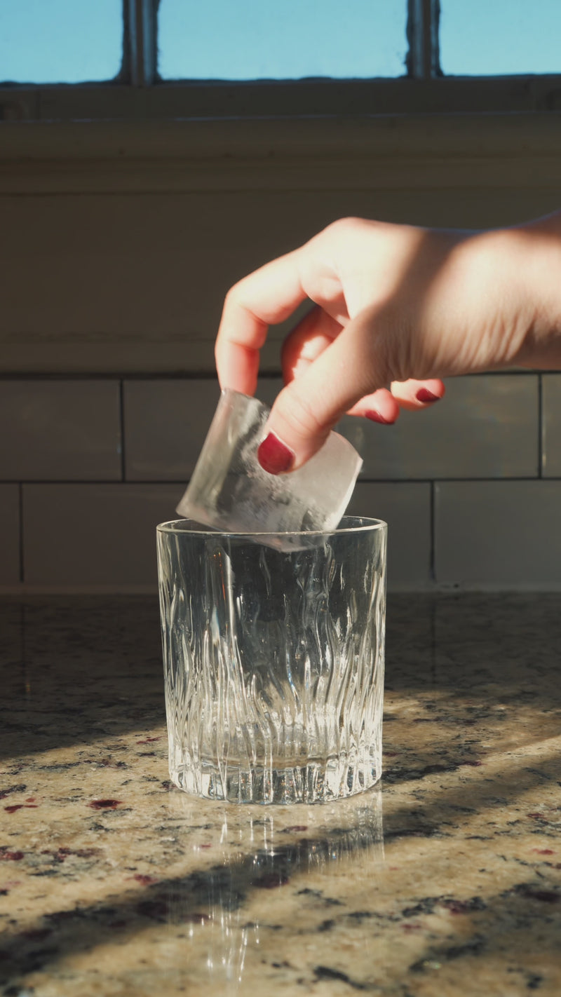 A clear, short rocks glass with a thick base is shown as ice is added. Lavender syrup is poured over the ice, then the glass is topped with tonic water, creating a lightly sparkling pour.