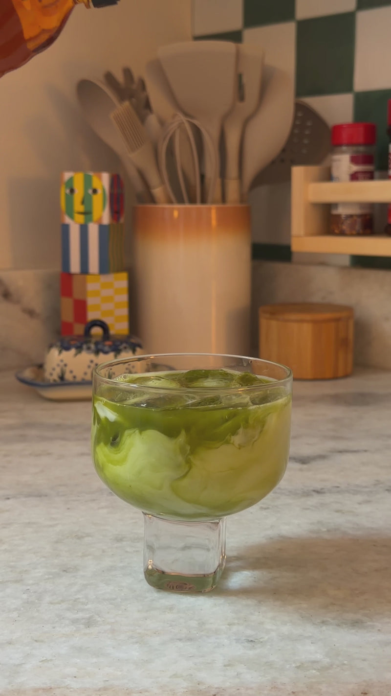 Maple syrup being poured into an iced matcha latte, creating a creamy swirl before stirring to combine.