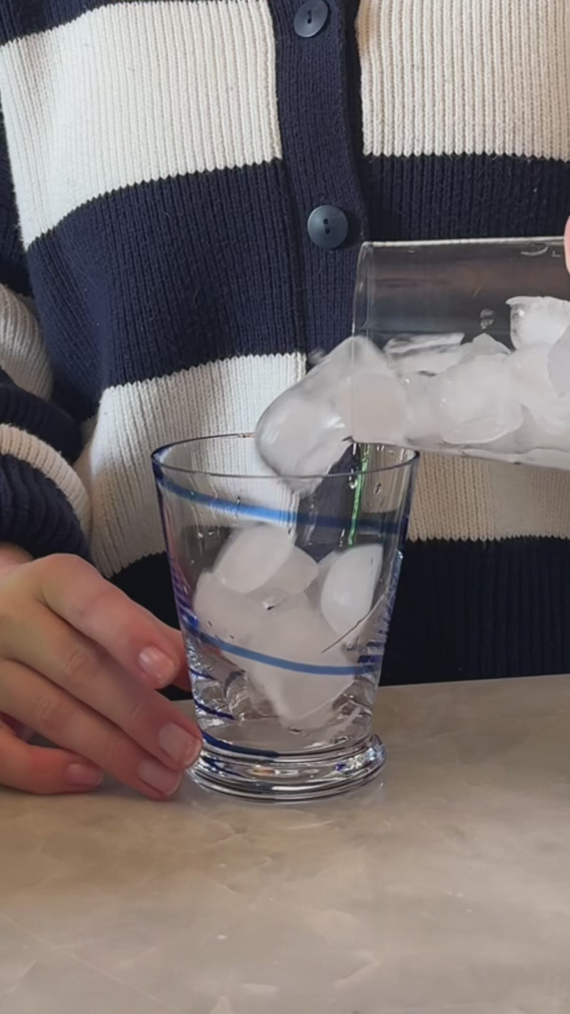 A clear, short rocks glass with a thick base sits on a neutral stone surface as ice fills the glass. Lemon-lime soda and soda water are poured over the ice, creating visible bubbles and a light, citrusy base.