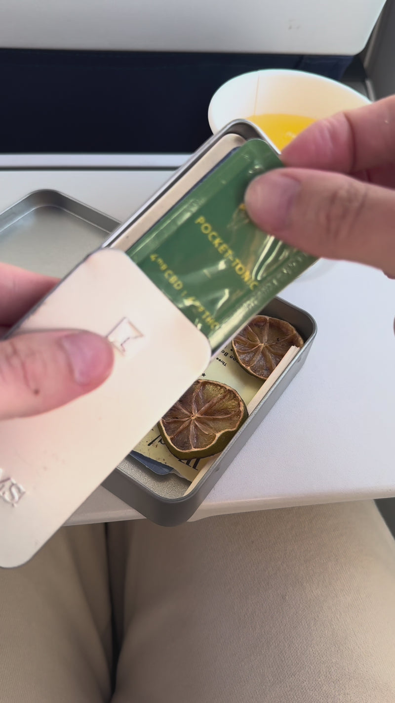 A Wims Lemon Basil Pocket-Tonic™ sachet is taken out of a Wims tin, snapped open, and added to the drink. A white straw stirs the mixture, followed by a dried lime wheel placed on top as the final garnish.