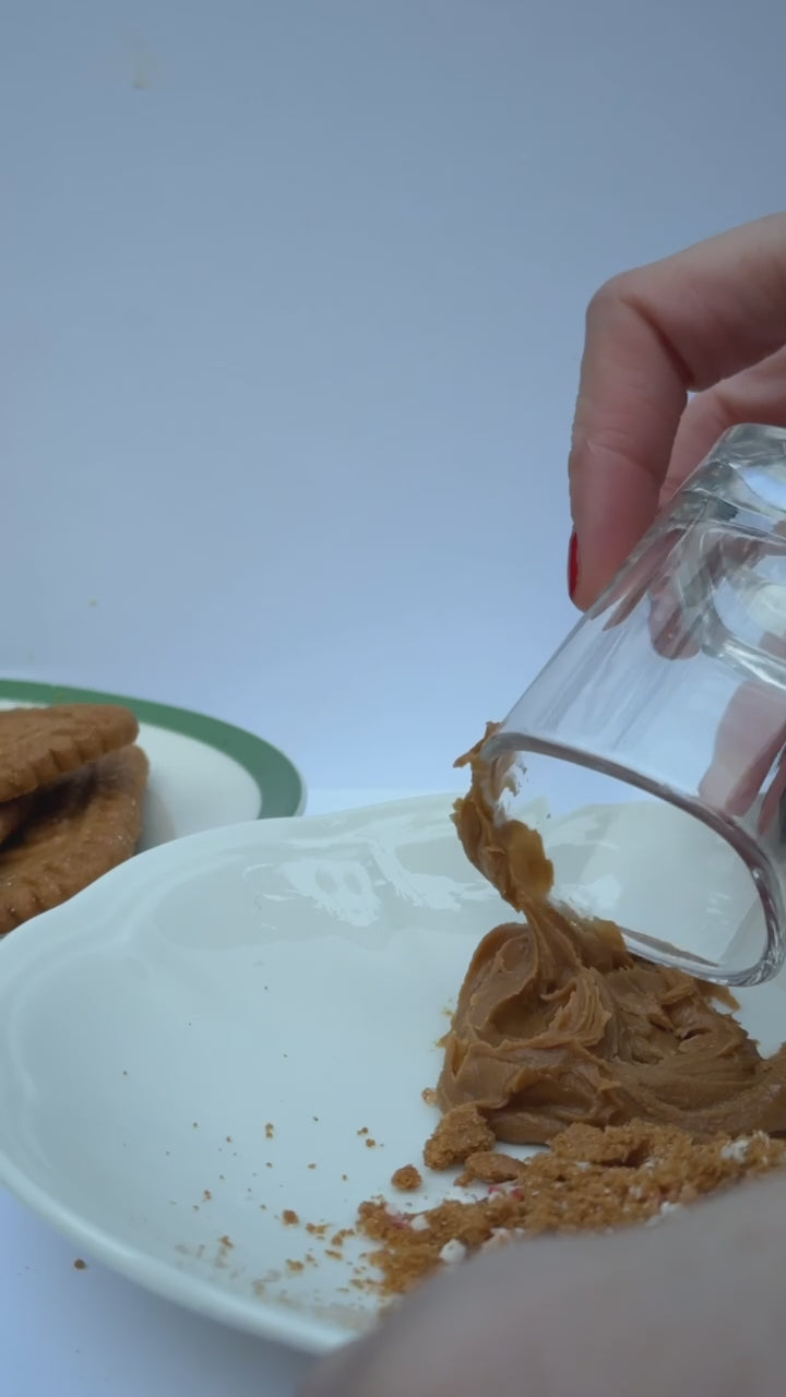  A hand dips the rim of a clear shot glass into cookie butter, then presses it into crushed cookies to create a coated edge.