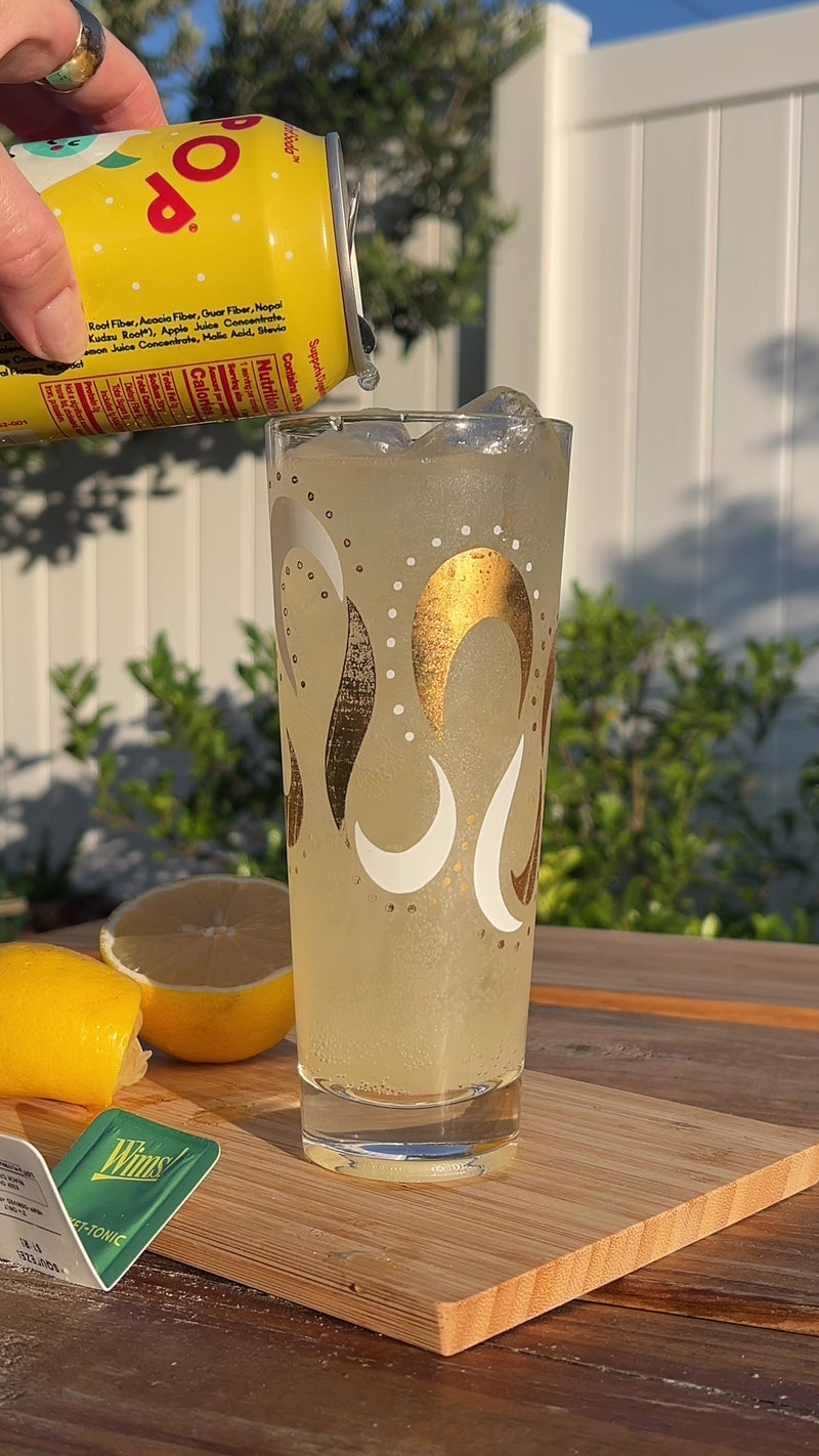 A tall glass of Sunny Cider Spritz being topped off with more Olipop Crisp Apple, then finished with a fresh basil sprig on top. Surrounding the glass are lemon slices, a Wims Lemon Basil Pocket-Tonic™ box, and an Olipop Crisp Apple can, creating a cozy fall vibe.