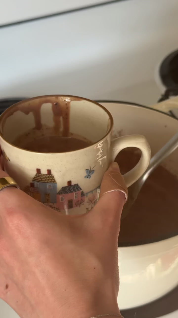A hand holds a ceramic mug decorated with small houses as hot chocolate is ladled in, showing how smooth and silky the mixture looks during the second step of making homemade hot chocolate.
