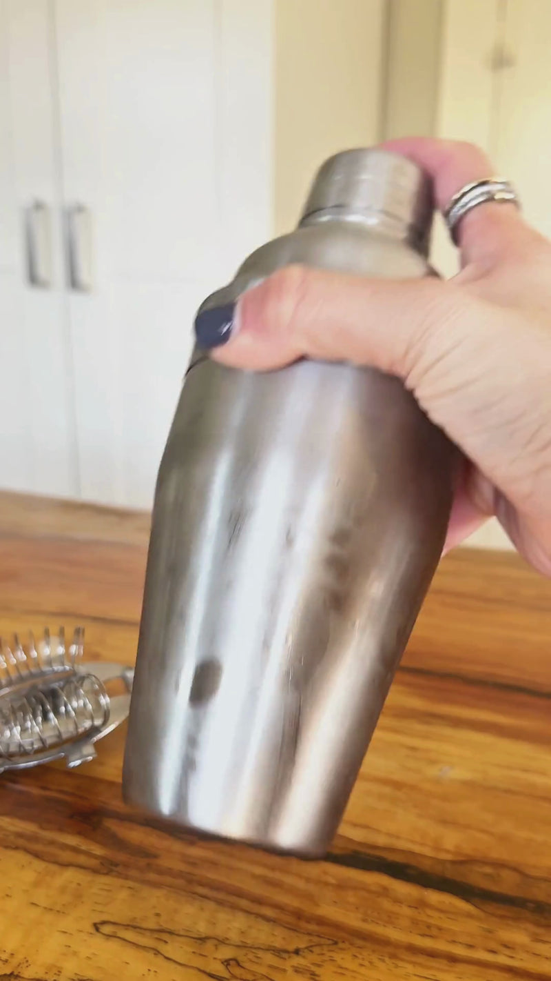 Stainless steel cocktail shaker being shaken, then a non-alcoholic Dirty Martini strained into a chilled martini glass and garnished with Wims-marinated olives.
