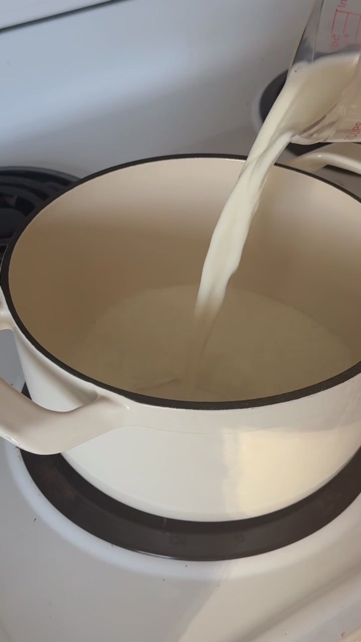 A hand pours milk into a white pot, then adds cocoa powder, dark chocolate, sweetened condensed milk, vanilla, a cinnamon stick, and a pinch of salt on a stovetop, beginning the first step of making homemade hot chocolate.