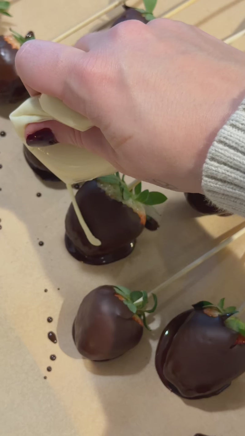 White chocolate drizzle being piped over chocolate-covered strawberries on parchment paper before chilling to set.