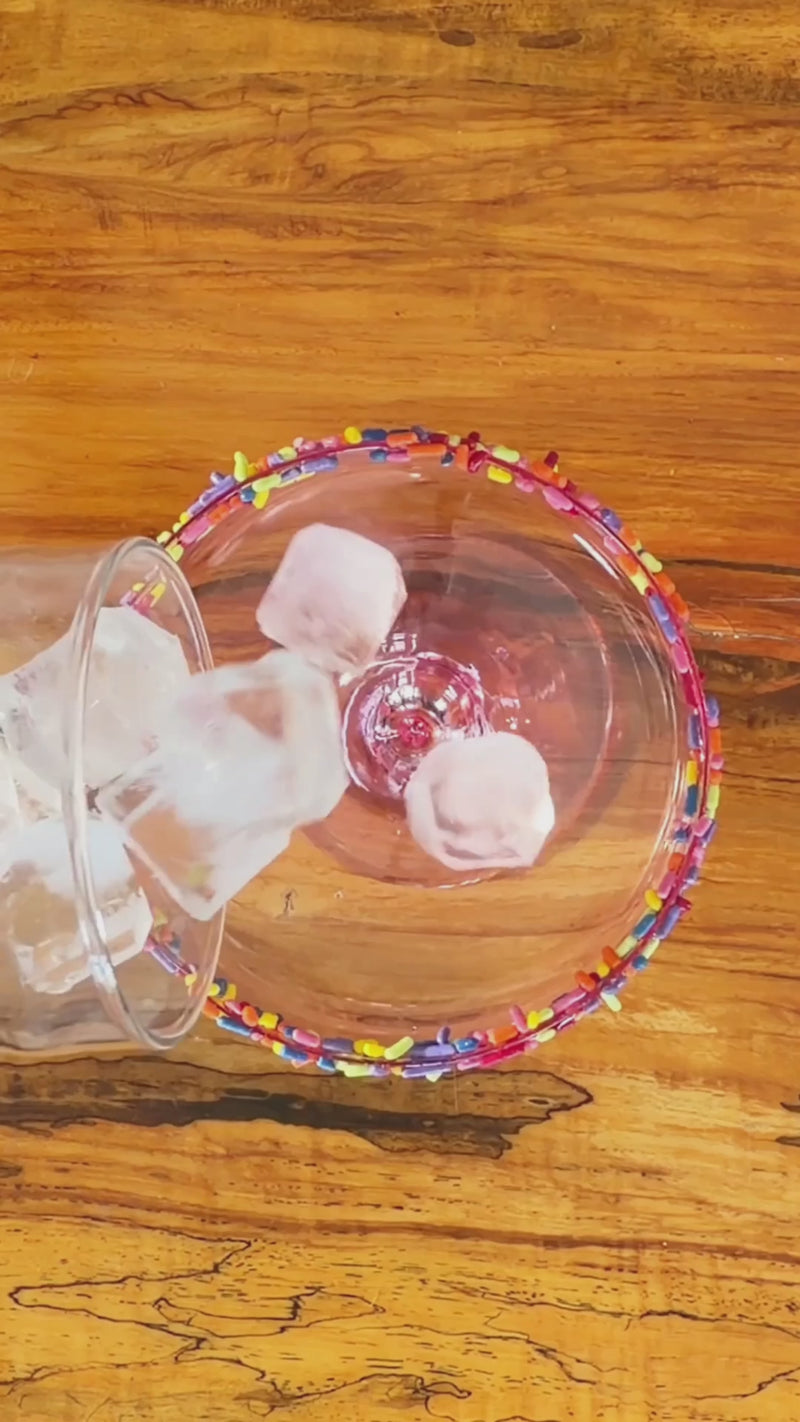 Coconut milk and watermelon juice being poured over ice into a rainbow-rimmed glass, creating a bright pink mocktail base.