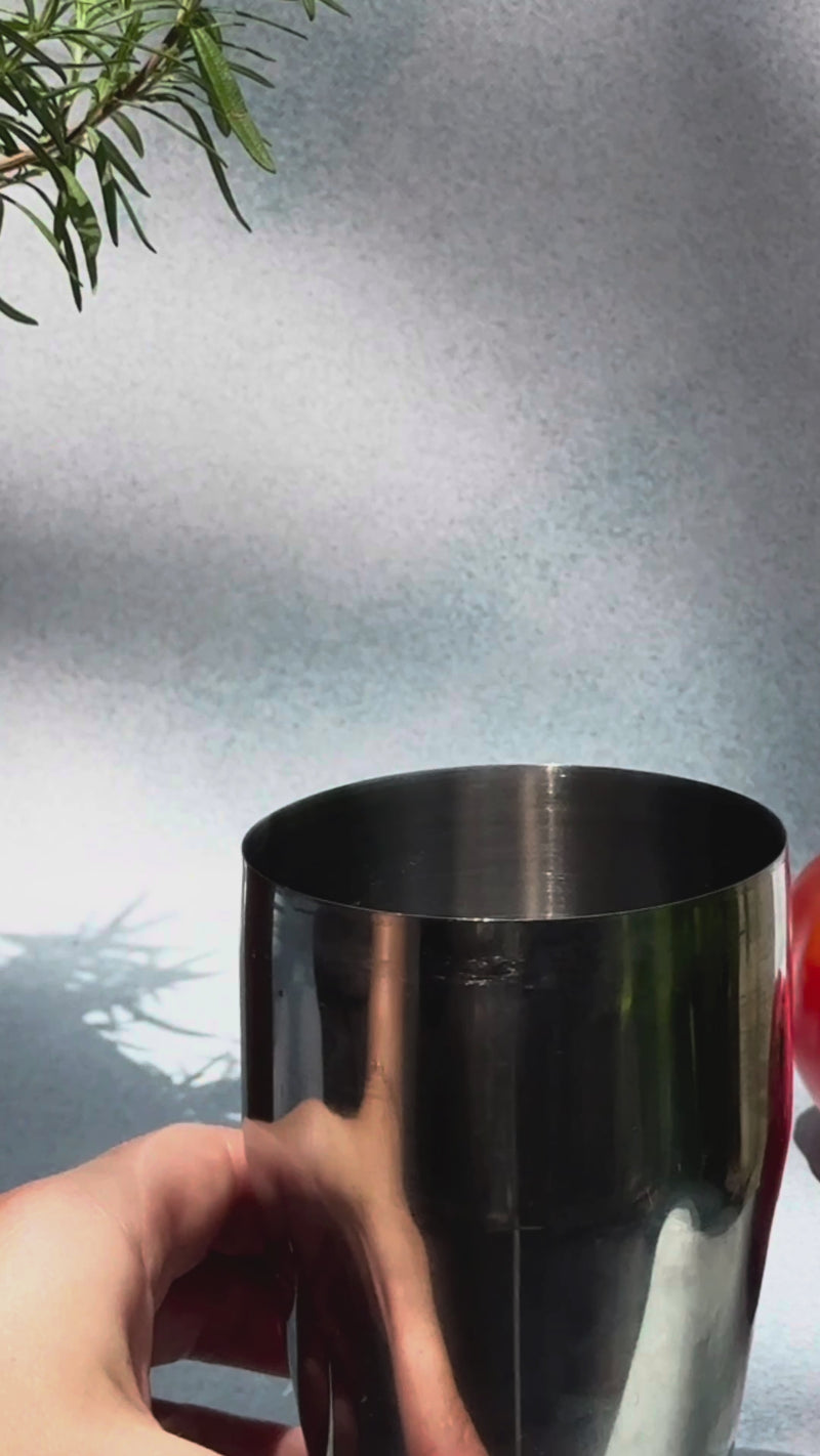 A hand adding ice cubes to a silver mixing cup, then pouring in tomato mix and tomato gin before shaking gently, with two fresh tomatoes in soft focus behind the cup.