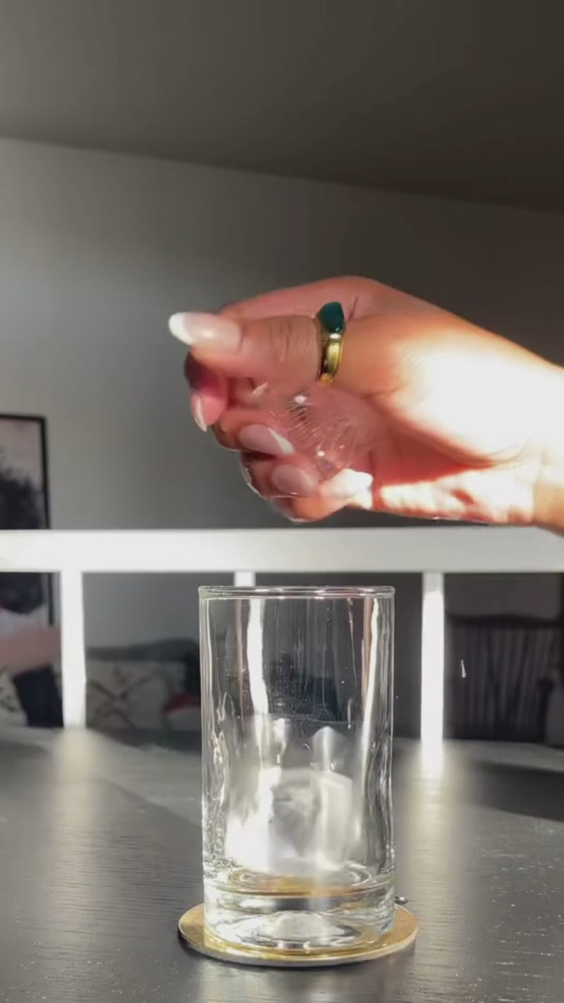 On a dark table with a cozy, modern home in the background, a tall glass is filled with a few ice cubes. Freshly grated ginger and lemon juice are added, creating a bright, tangy base at the bottom of the glass.