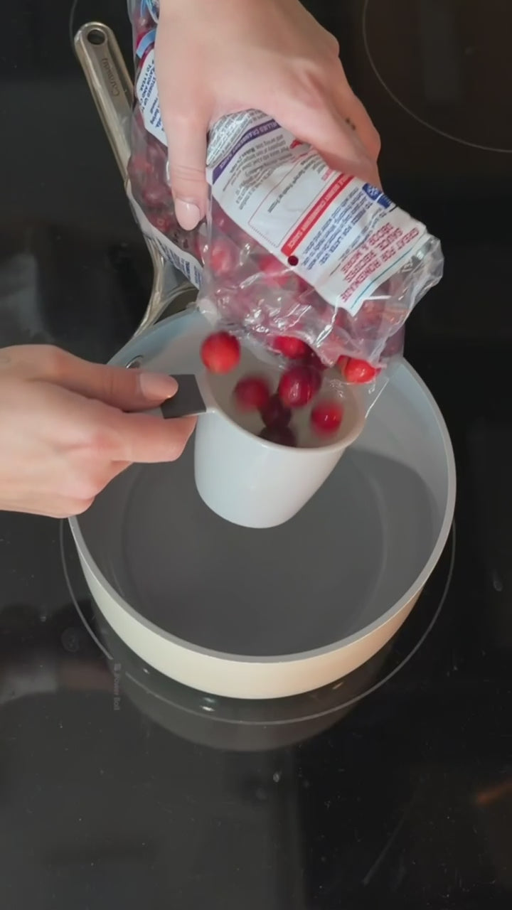 Hand adding cranberries, orange juice, sugar, and lime juice into a small saucepan on the stovetop, stirring as the ingredients combine to start a cranberry citrus syrup.