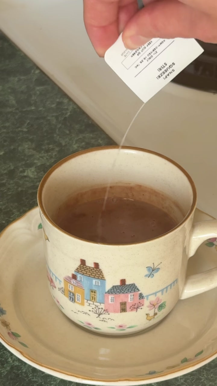 A hand snaps open a Wims Unflavored Pocket-Tonic™ and pours it into the same ceramic mug of hot chocolate, showing the final step of stirring in the Wims sachet before serving.