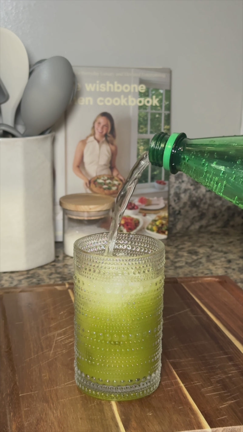 A finished green cucumber drink in a textured glass filled with ice, topped with soda water for a light, bubbly finish, as a hand places a fresh lime wedge on the rim, with extra lime slices on a wooden cutting board and a cozy kitchen setup in the background.
