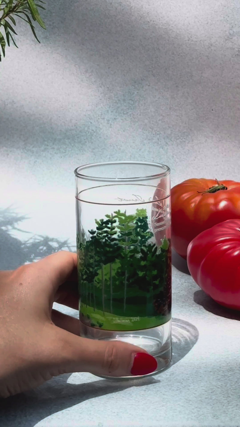 A hand adding ice to a tree-illustrated Collins glass, topping with sparkling volcanic water, snapping in a Wims Lemon Basil Pocket-Tonic™ (4mg THC, 4mg CBD), and stirring with a toothpick garnished with a cherry tomato and fresh green leaf. Two ripe tomatoes rest beside the glass in the background.
