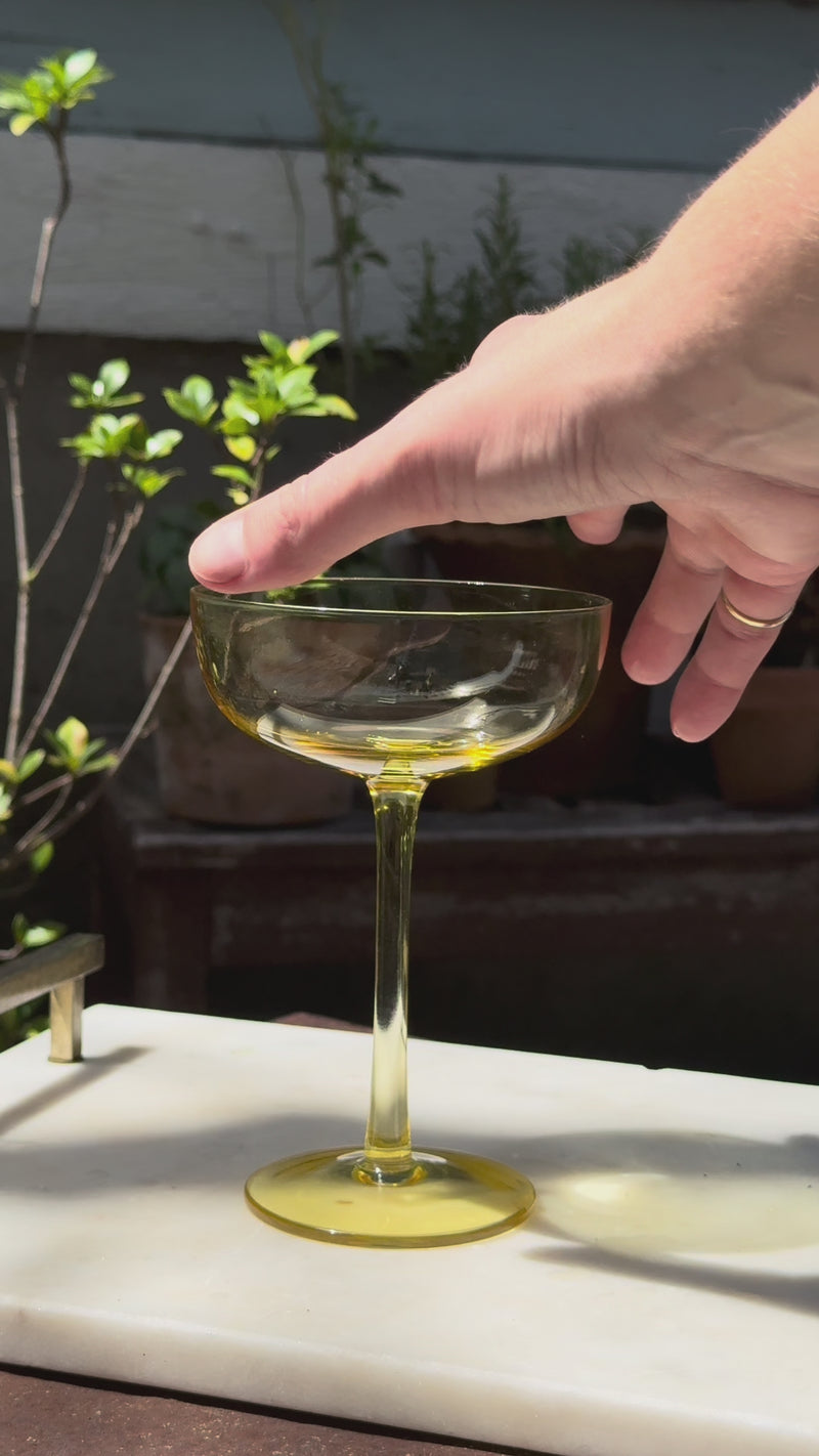 Golden-yellow Kitchen Garden cocktail being poured into a matching yellow-stemmed glass, later topped with soda water and garnished with fresh rosemary. Sunlit garden plants in the background.